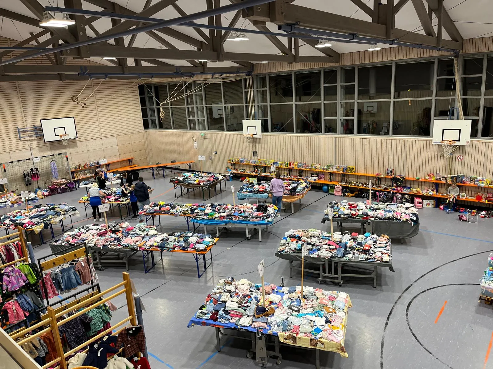 Blick von der Empore in die Turnhalle der Grundschule Fronberg: Reihen von Tischen mit nach Größen sortierten Kindersachen, Kleiderständer an der Wand, einzelne Helferinnen und Helfer beim Aufbau